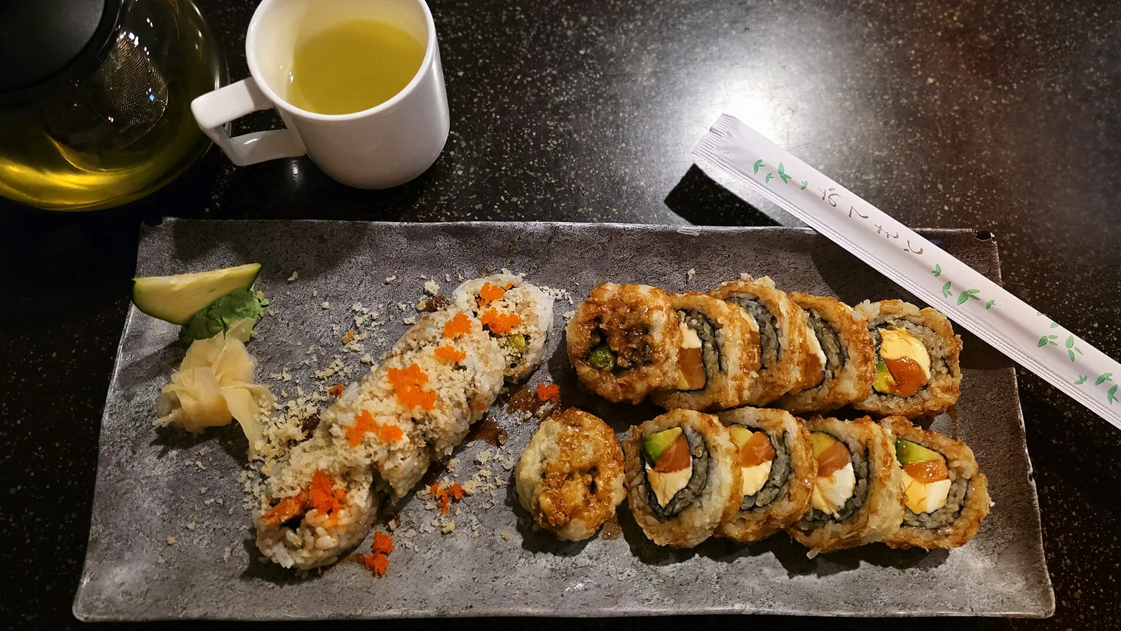 Two sushi rolls on a slate plate with ginger and wasabi, beside a cup of green tea.